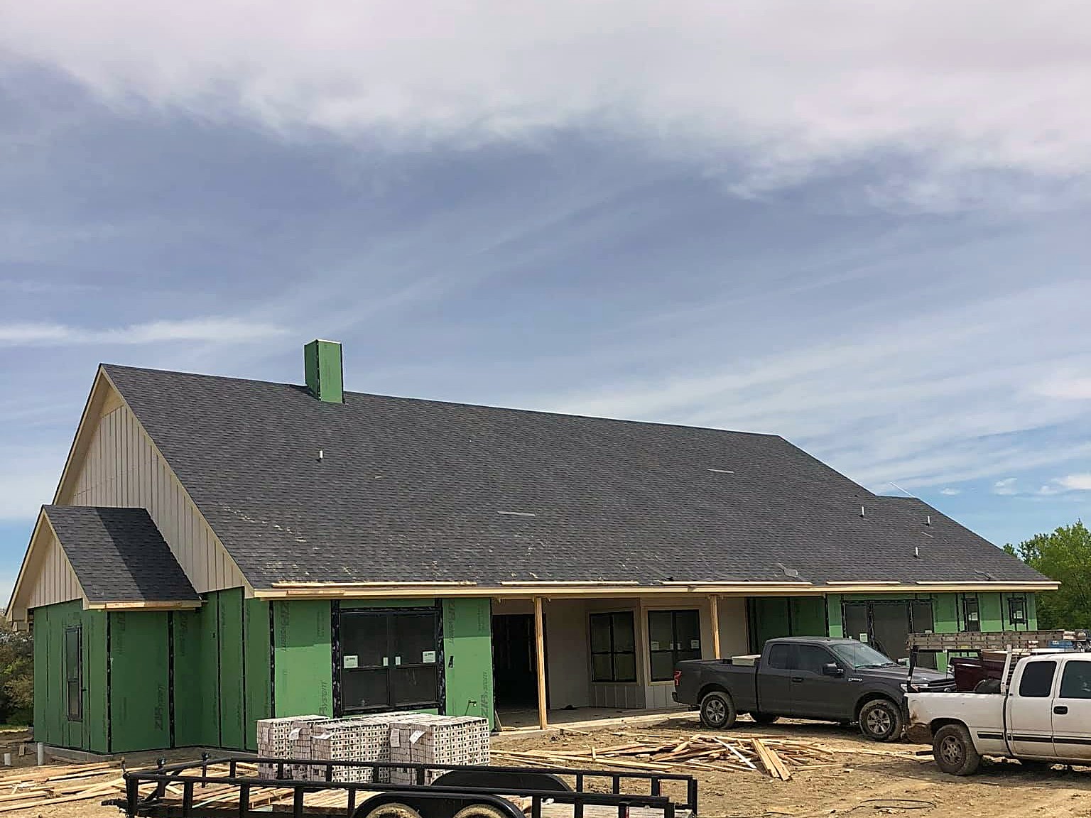 A long house with a finished dark shingle roof, green sheathing below it and pickups in the yard