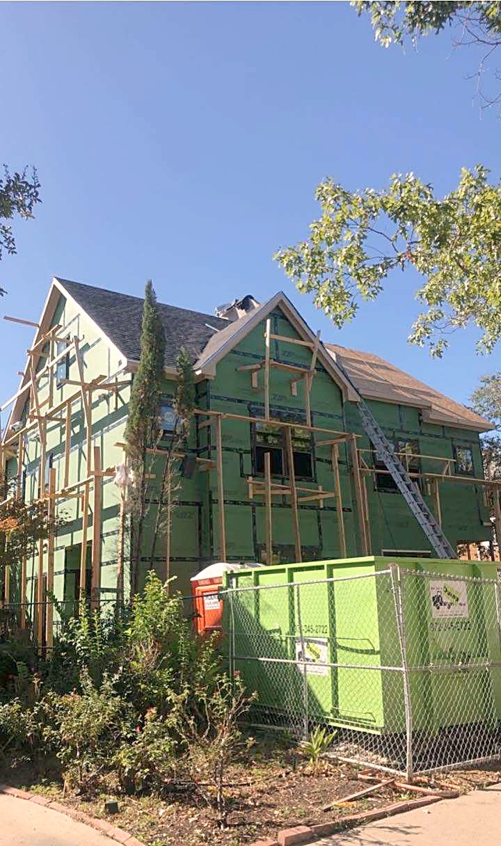 A two-story house wrapped in green sheathing with a ladder up to the roof and a dumpster in the yard